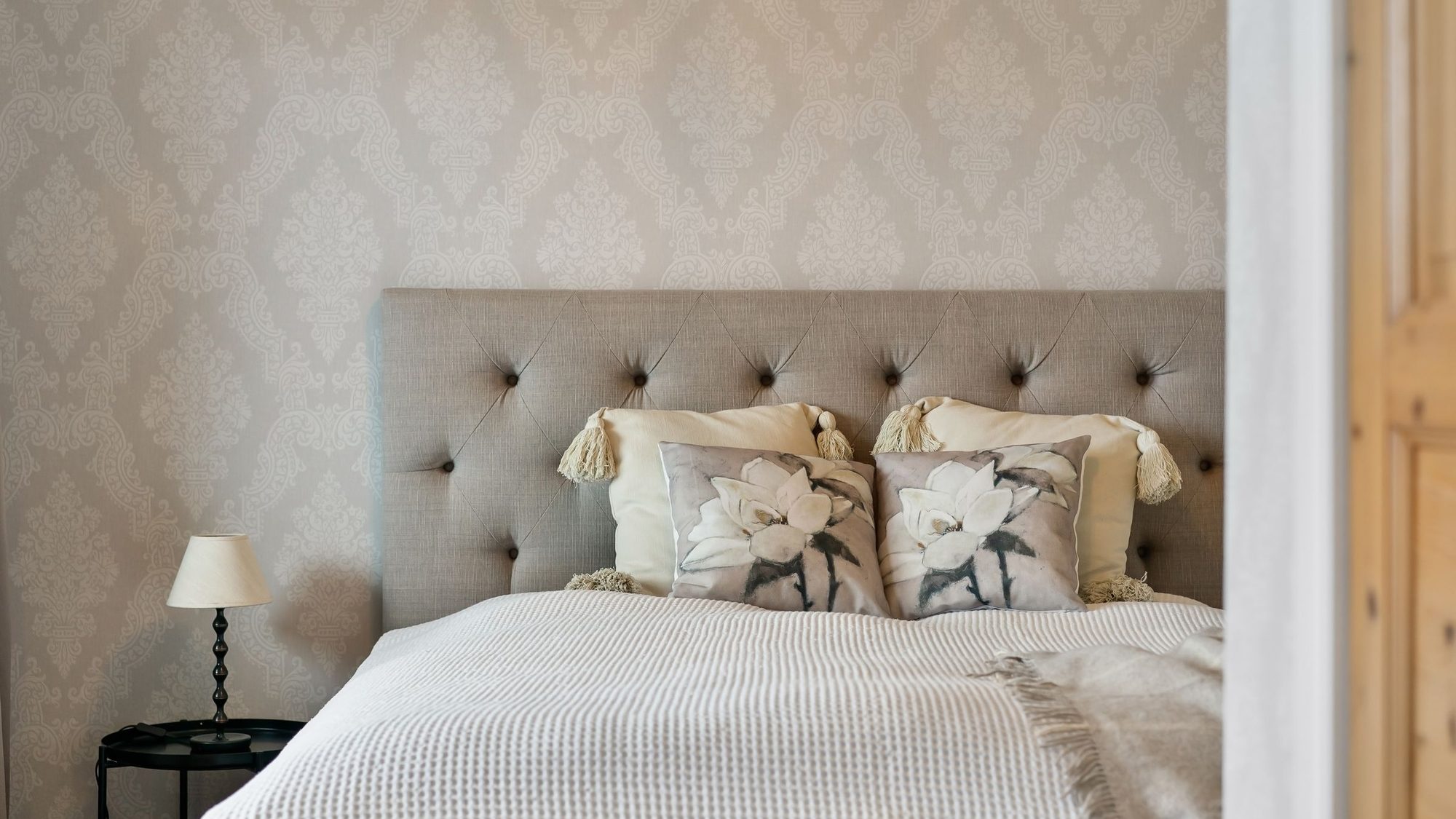 An elegant residential bedroom finished with soft damask wallpaper behind an upholstered headboard